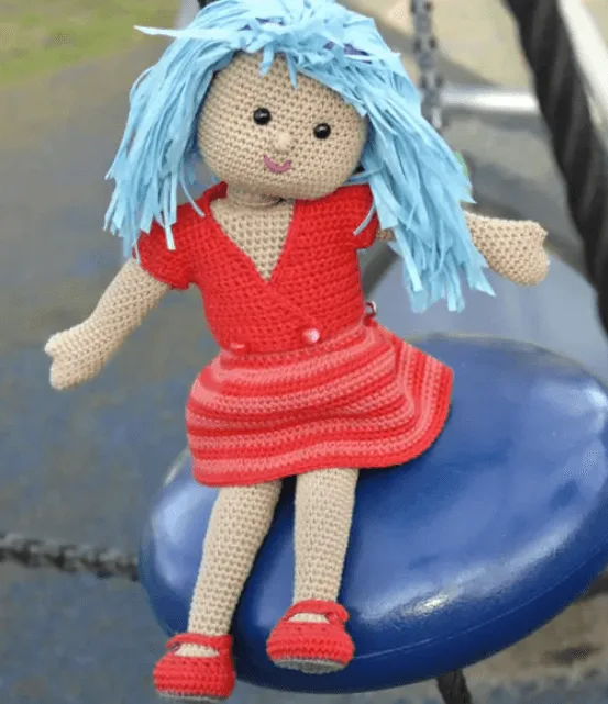 Poup&eacute;e au crochet avec une robe rouge et des cheveux bleus, assise sur un jeu ext&eacute;rieur, cr&eacute;&eacute;e &agrave; partir du livre "Les poup&eacute;es de Tyhia au crochet".
