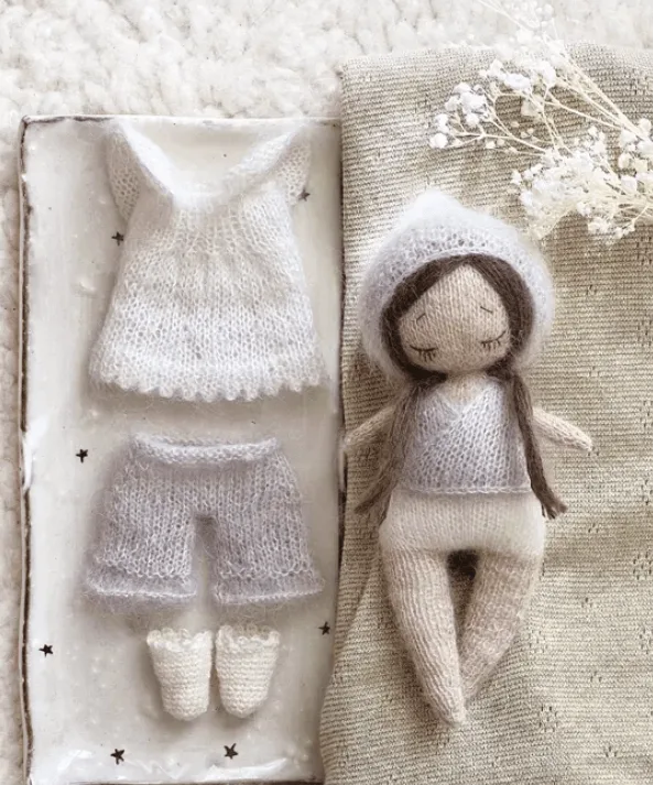 Poup&eacute;e Pom'ette en laine accompagn&eacute;e de sa garde-robe tricot&eacute;e, comprenant une robe, un pantalon et des chaussons, expos&eacute;e dans un d&eacute;cor doux et minimaliste.
